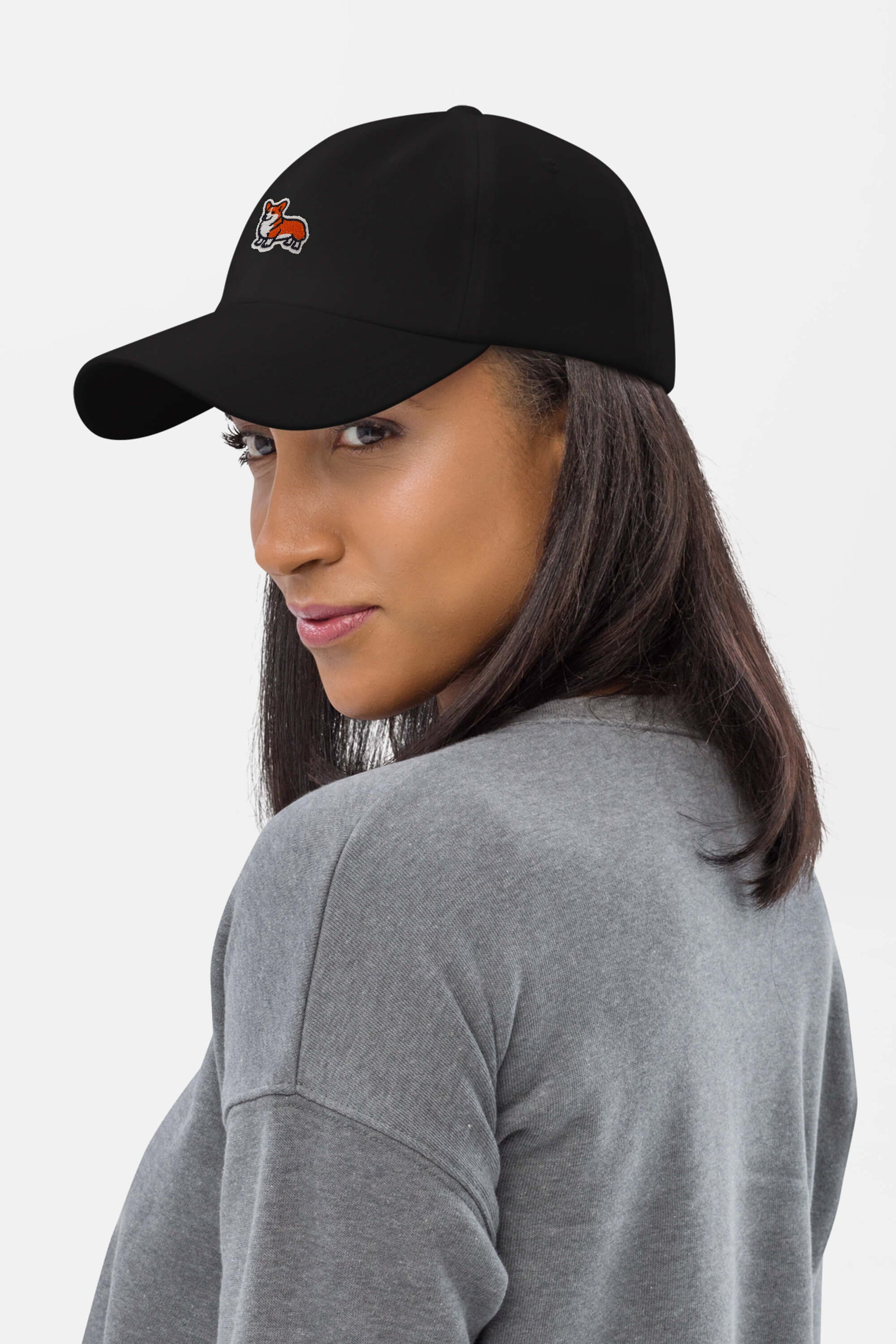 A woman with straight brown hair models the Iconic Corgi Cap by Dogloverclothing.com—a black hat featuring a small fox logo—paired with a gray sweatshirt, glancing over her shoulder against a plain white background.