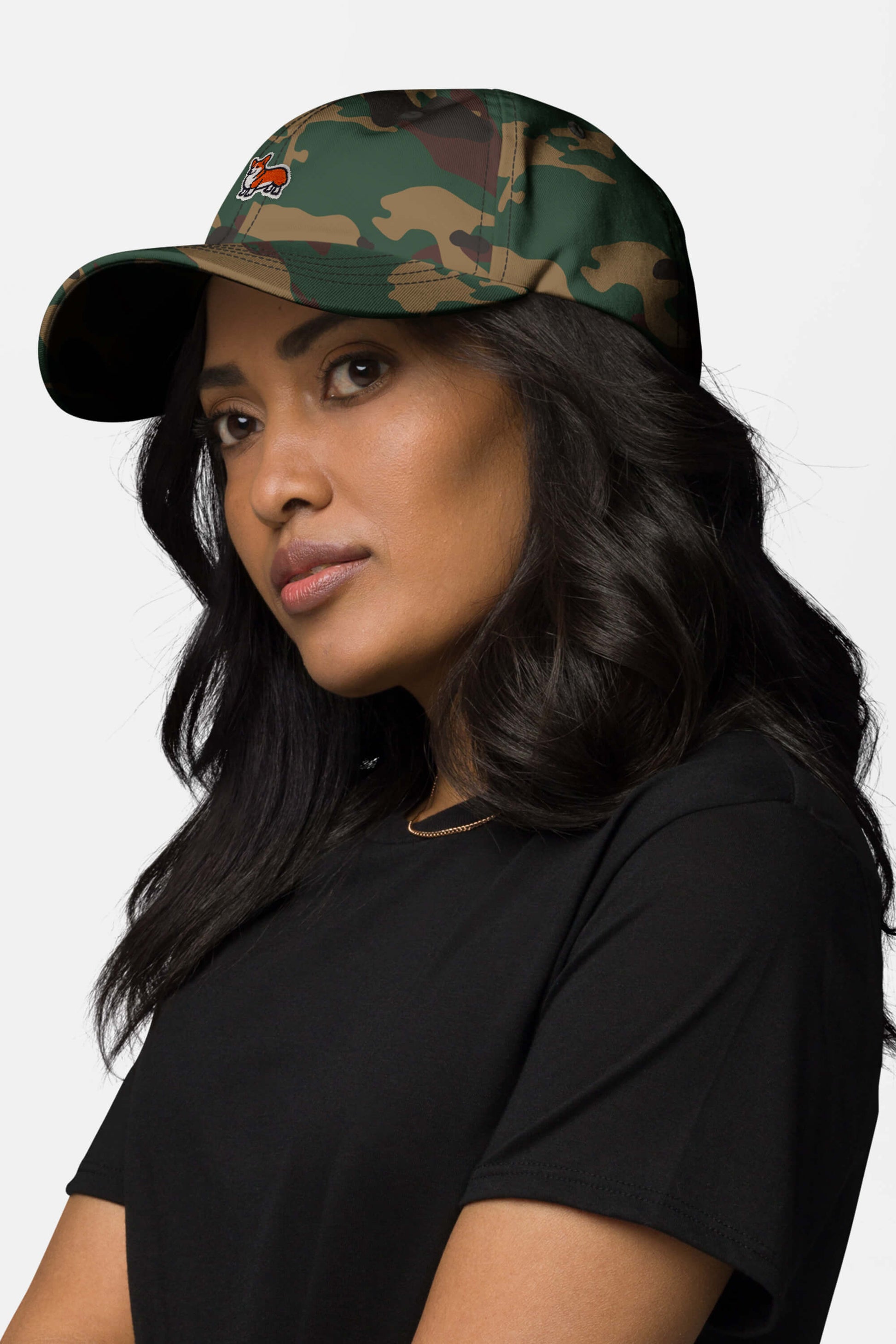 A woman with long dark hair wears the Iconic Corgi Cap from Dogloverclothing.com and a black t-shirt, glancing to the side against a plain light background, showcasing Corgi-inspired style in her casual look.