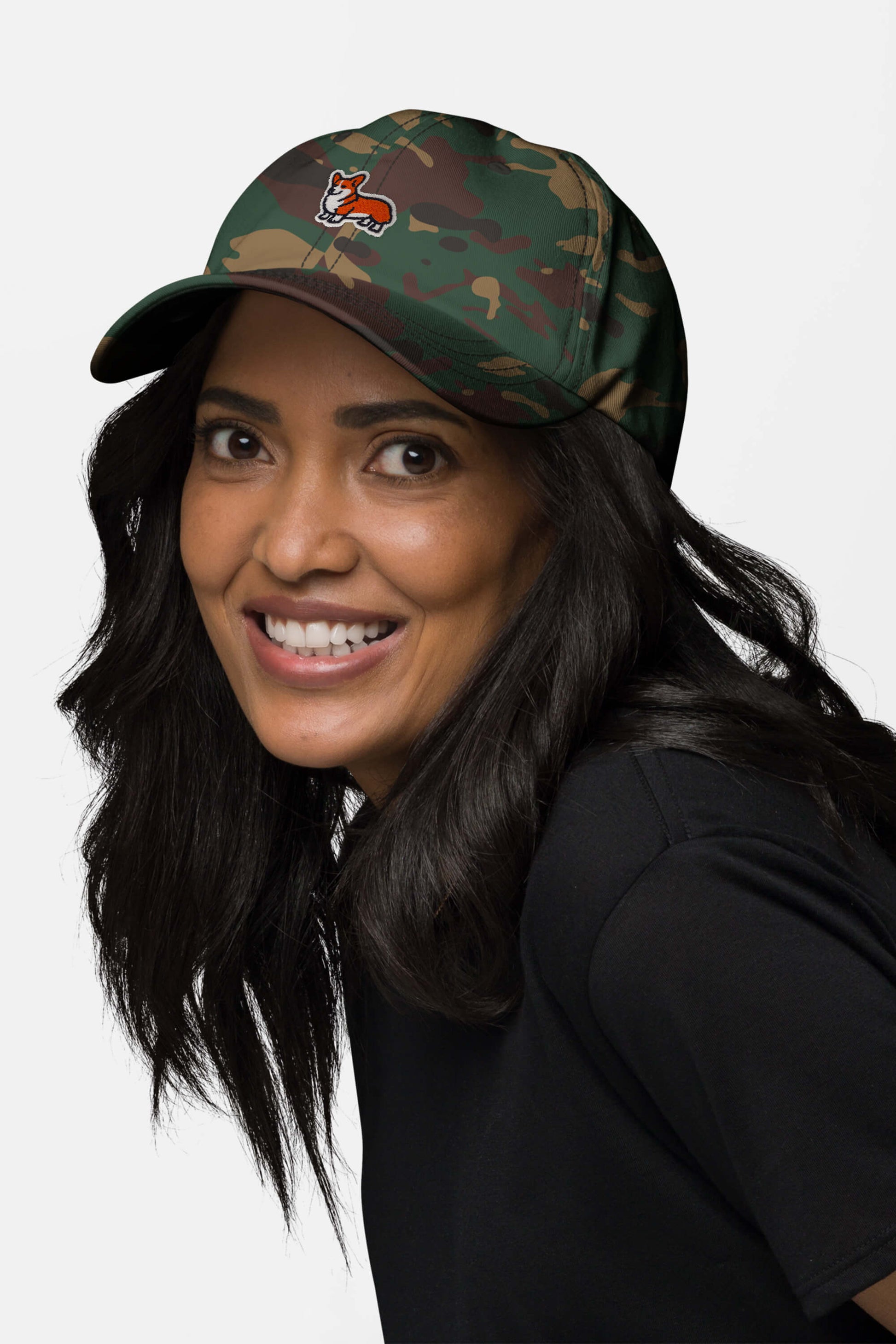 A smiling woman with long dark hair and a black shirt models the Iconic Corgi Cap by Dogloverclothing.com, a green camouflage hat with a small embroidered fox logo, set against a plain light background.