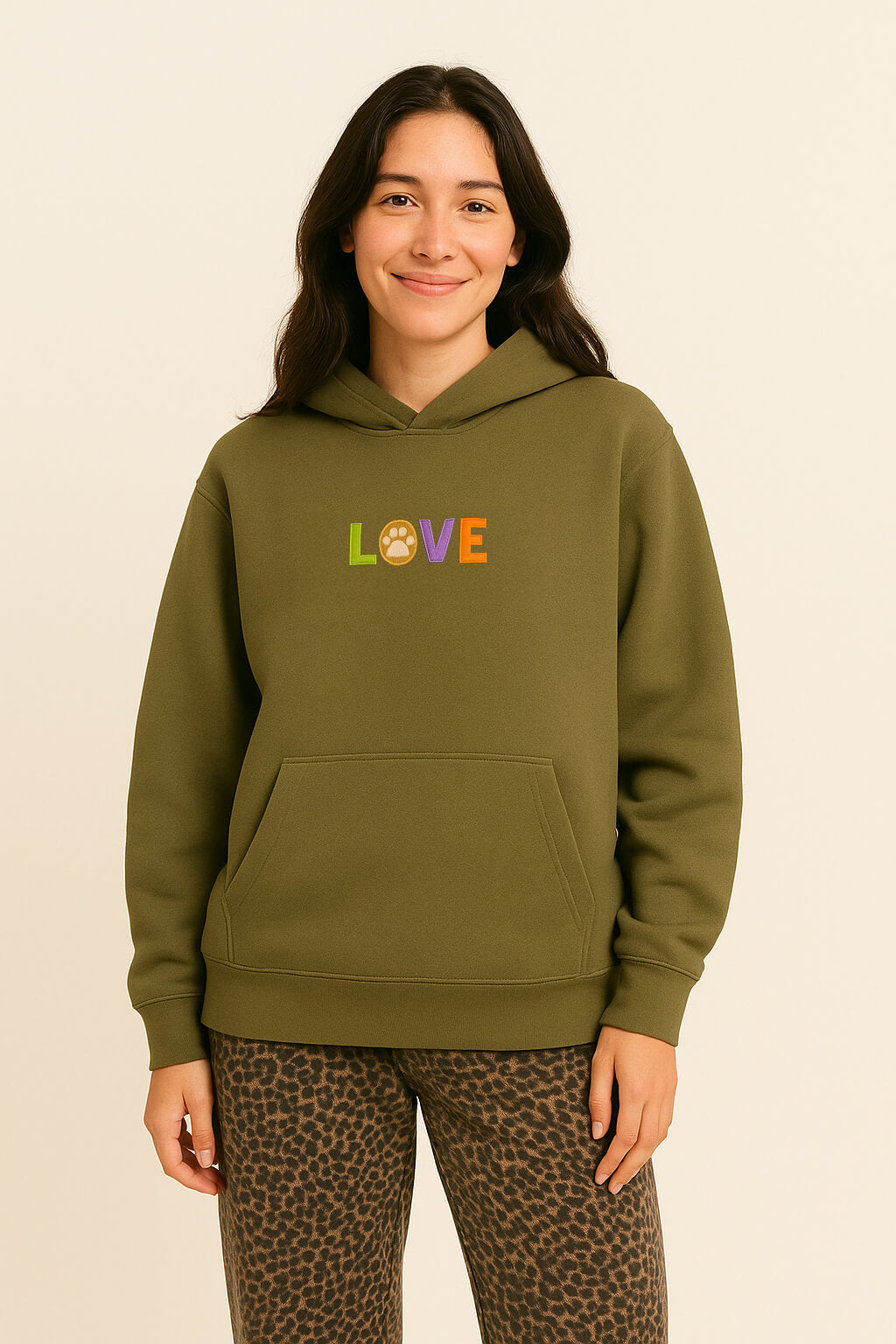 A smiling woman with long dark hair models the Super Soft Dog Love Hoodie by Dogloverclothing.com—an olive green top with an embroidered LOVE design featuring a paw print as the O—paired with leopard print pants against a beige background.