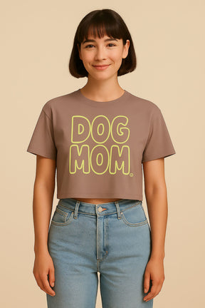 A smiling person wears the Oversized Dog Mom Crop Top by Dogloverclothing.com, featuring bold yellow DOG MOM lettering, styled with high-waisted light blue jeans against a plain beige background.
