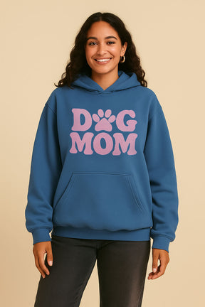 A smiling person wears the Dogloverclothing.com Dog Mom Hoodie, an ultra-soft blue hoodie with large pink “Dog Mom” text and a playful paw-print, standing against a plain beige background.