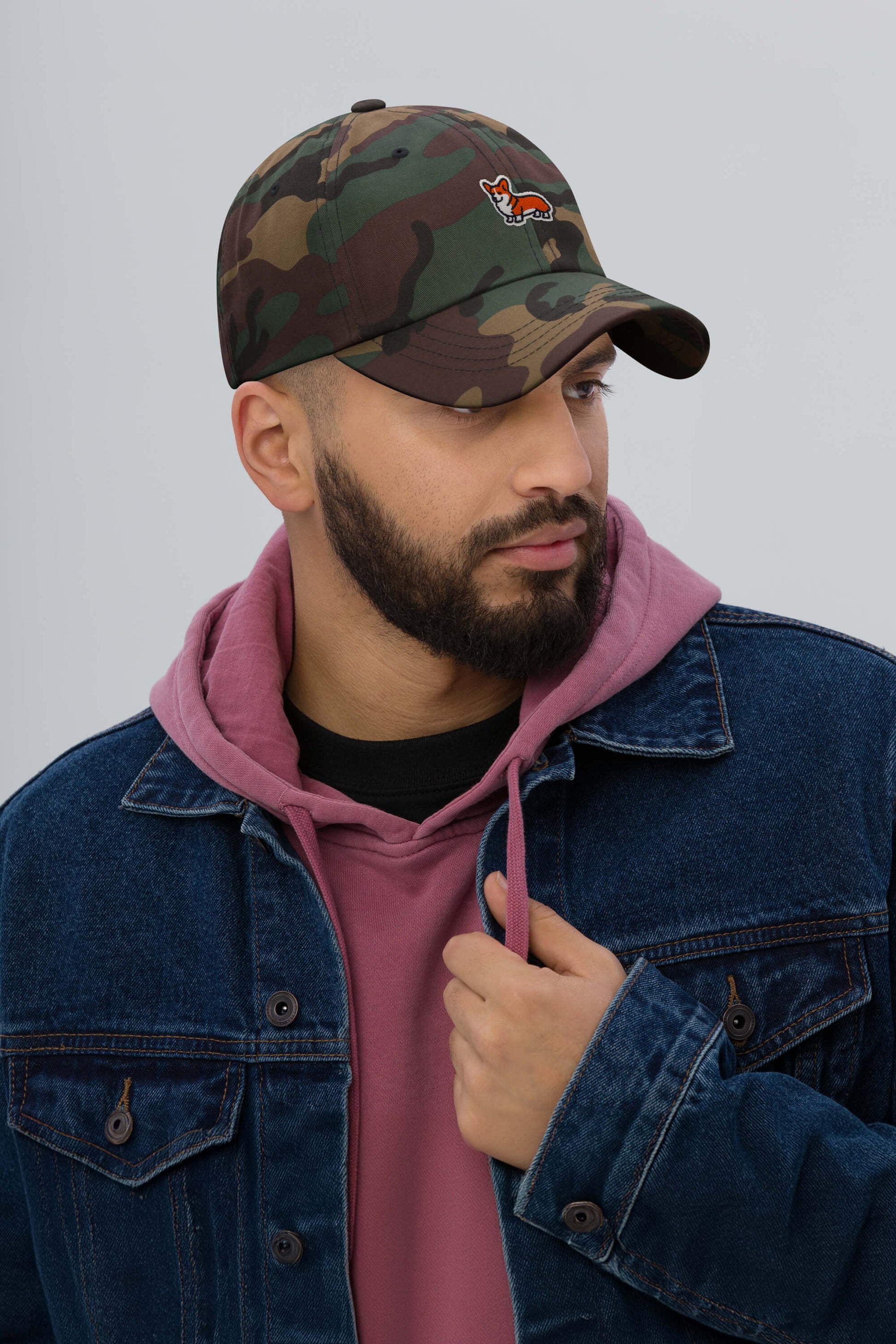 A bearded man wears the Iconic Corgi Cap from Dogloverclothing.com—a camouflage hat with corgi design—paired with a pink hoodie and blue denim jacket, gazing sideways against a plain light background.