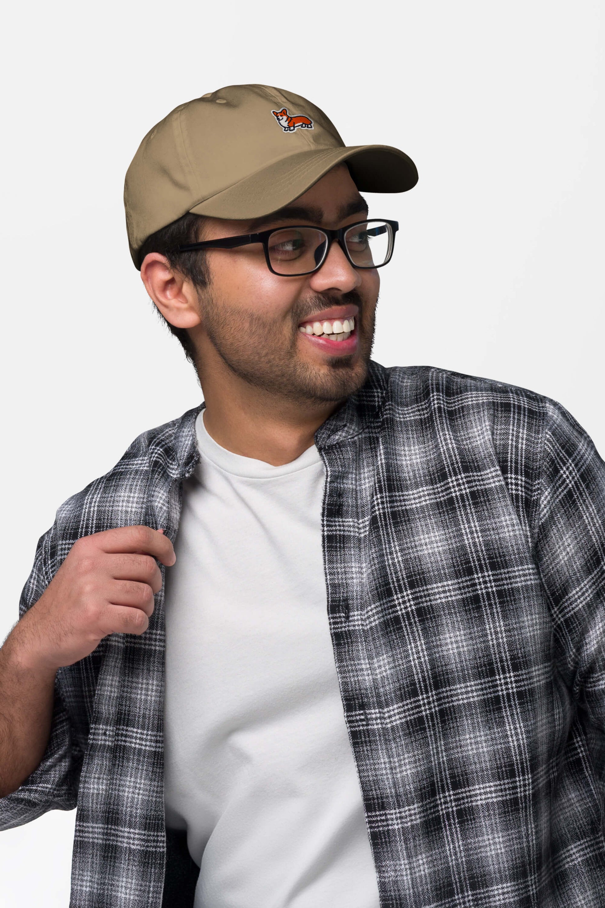 A man in glasses, a white tee, and a black-and-white plaid shirt smiles to the side while wearing the Iconic Corgi Cap from Dogloverclothing.com, showing off its small embroidered fox against a plain light background.