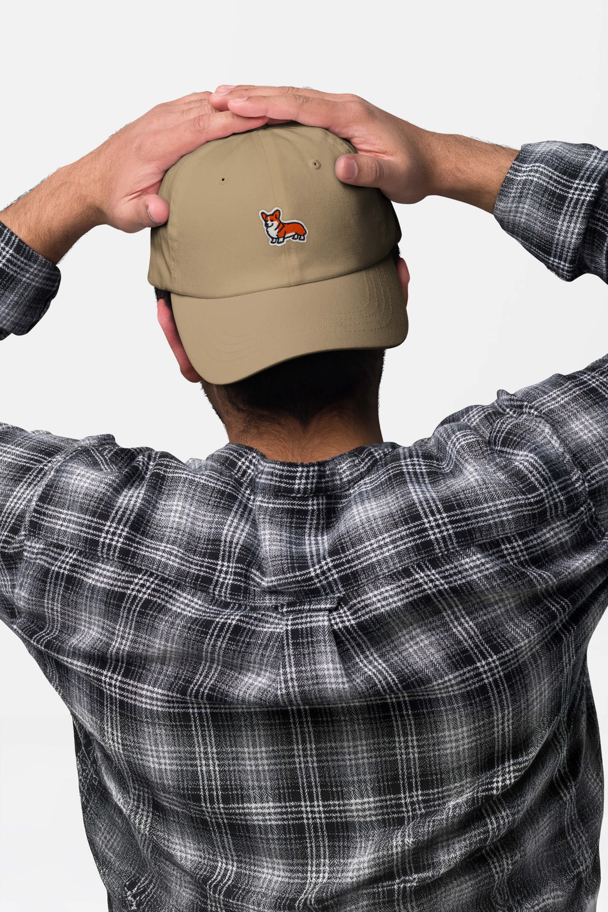 A person in a Dogloverclothing.com Iconic Corgi Cap—tan with a small fox patch—pairs it with a black-and-white plaid shirt, standing hands behind head to show off this cute, casual accessory from the Dogloverclothing.com collection.