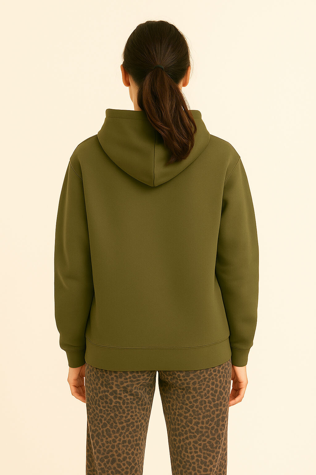 A person with long dark hair in a ponytail stands with their back to the camera, wearing Dogloverclothing.com’s Super Soft Dog Love Hoodie in olive green and brown leopard print pants, against a light beige background.