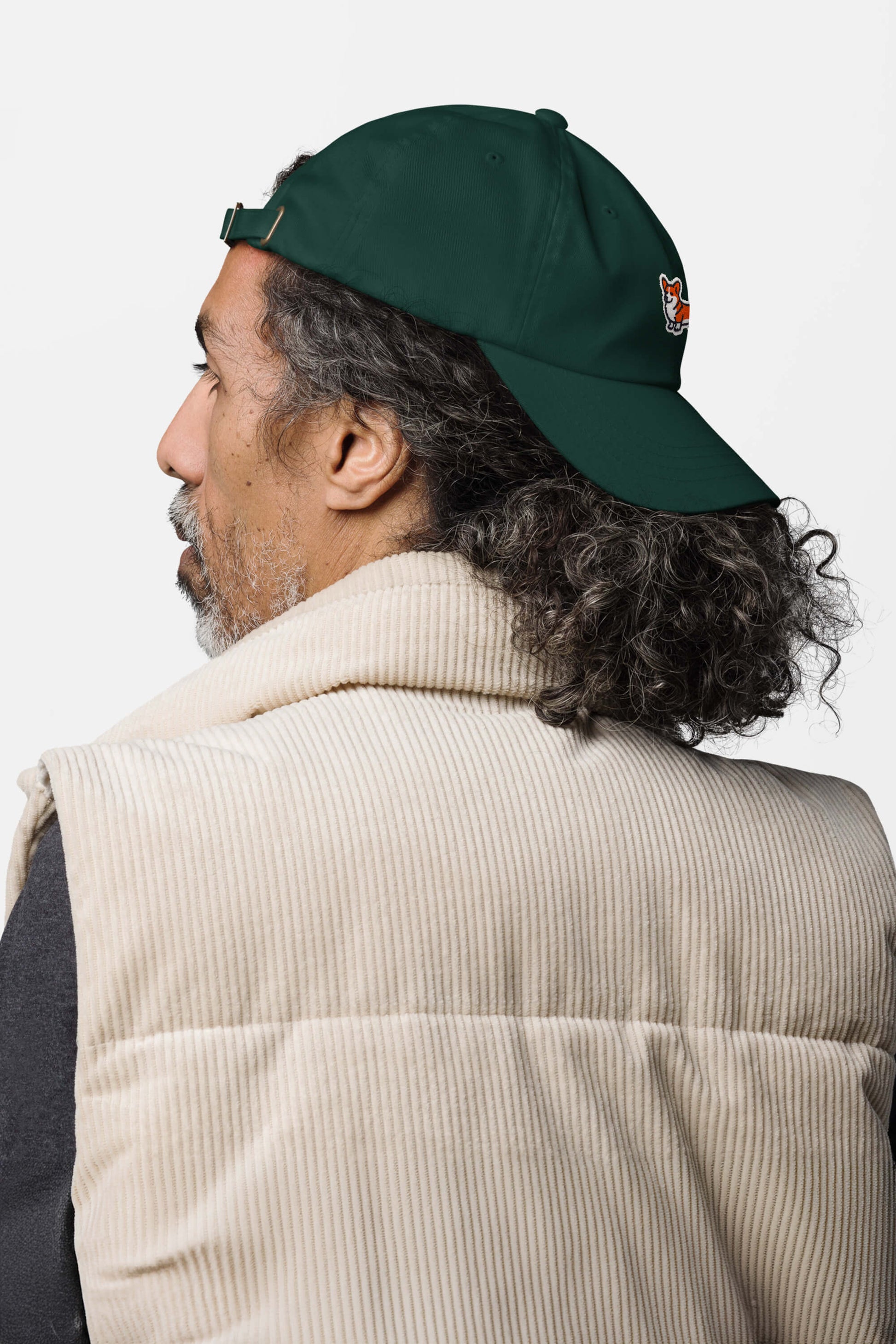 A man with curly gray hair wears a dark green Iconic Corgi Cap from Dogloverclothing.com backwards and a beige corduroy vest, shown from behind against a plain white background.