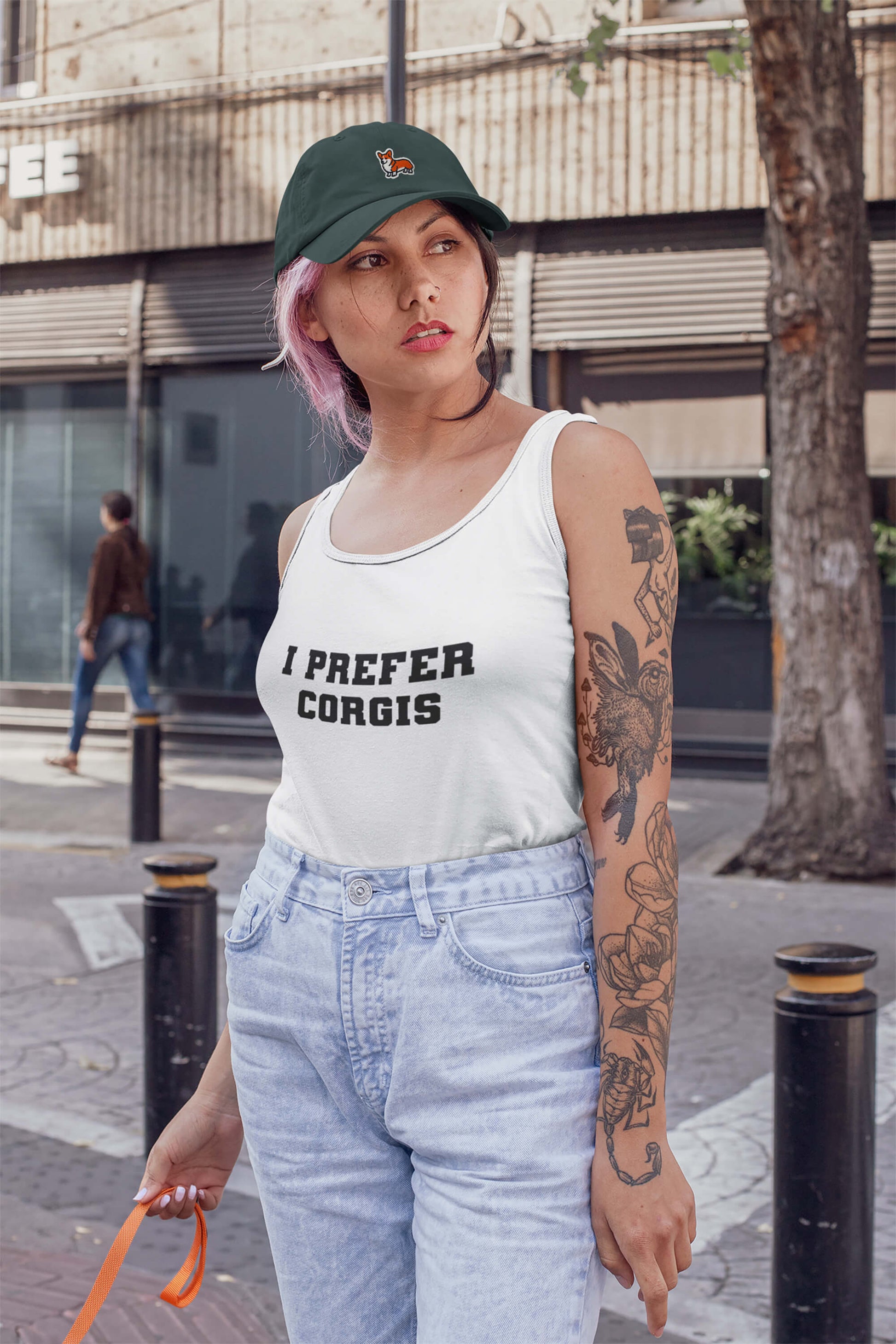 A woman with pink hair and tattoos wears a white I PREFER CORGIS tank top and the Dogloverclothing.com Iconic Corgi Cap. She stands on a city sidewalk, holding an orange leash, with buildings visible in the background.