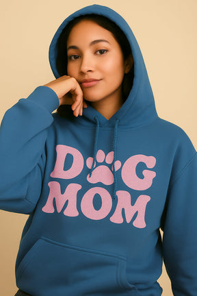 A woman models the Dogloverclothing.com Dog Mom Hoodie, featuring large pink DOG MOM lettering and a playful paw-print design, as she poses with her hand on her chin against a beige background.