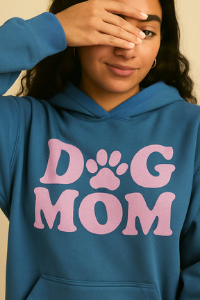 A woman wears the ultra-soft Dog Mom Hoodie from Dogloverclothing.com, featuring large pink DOG MOM letters with a paw-print O. She smiles softly, playfully covering one eye with her hand.