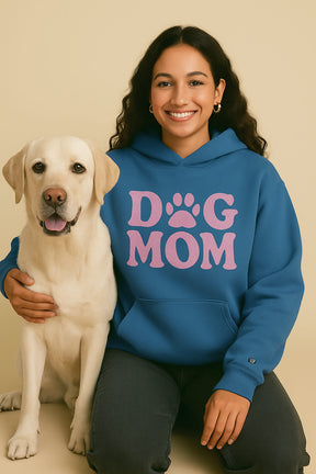 A smiling woman in a blue Dog Mom Hoodie from Dogloverclothing.com, featuring “DOG MOM” and a paw-print design, sits with a happy yellow Labrador Retriever against a beige background, her arm gently around the dog.