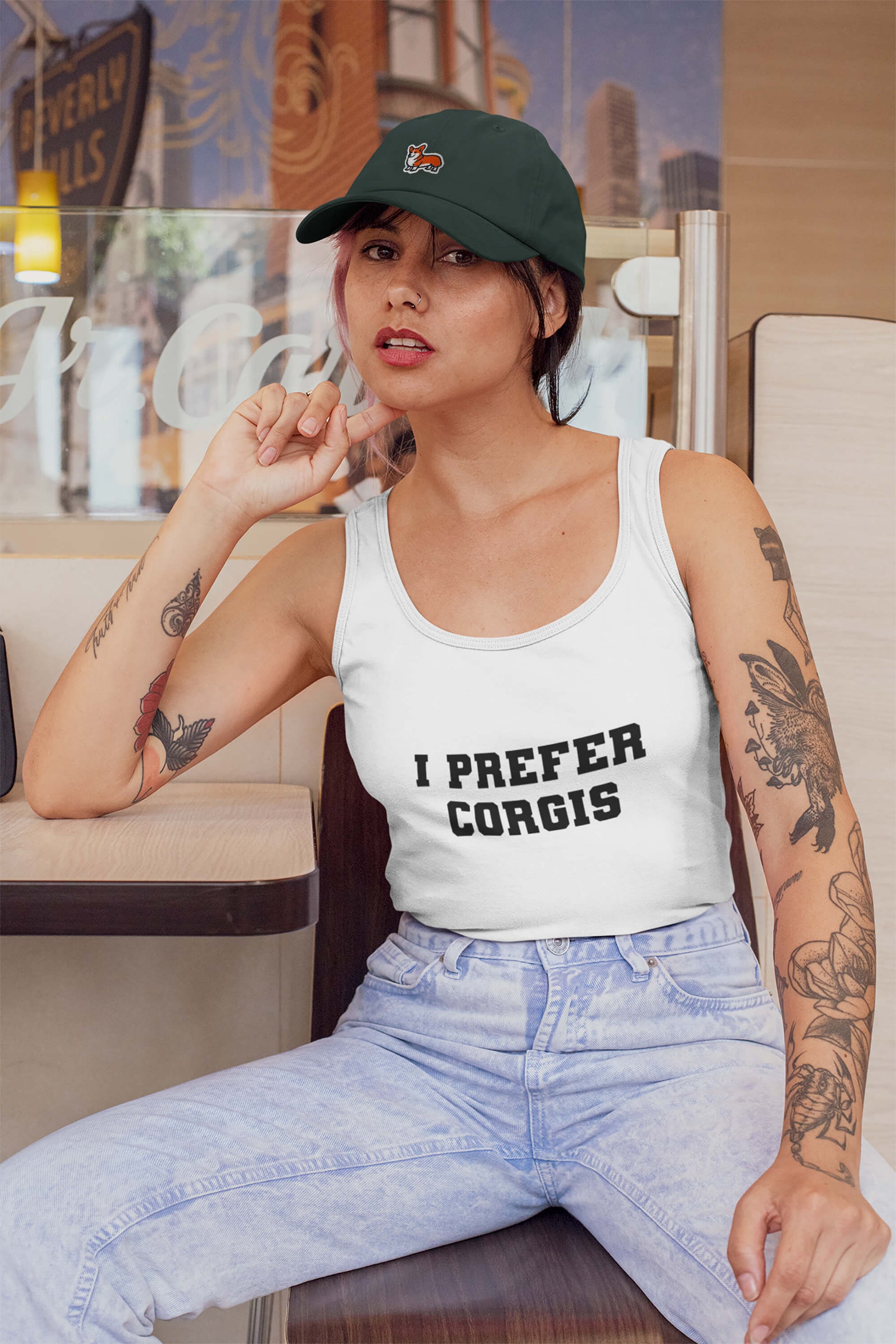 A tattooed woman in a green cap sits calmly in a café booth, wearing Dogloverclothing.coms Iconic I Prefer Corgis Super Tank Top, and poses with one hand touching her hat.