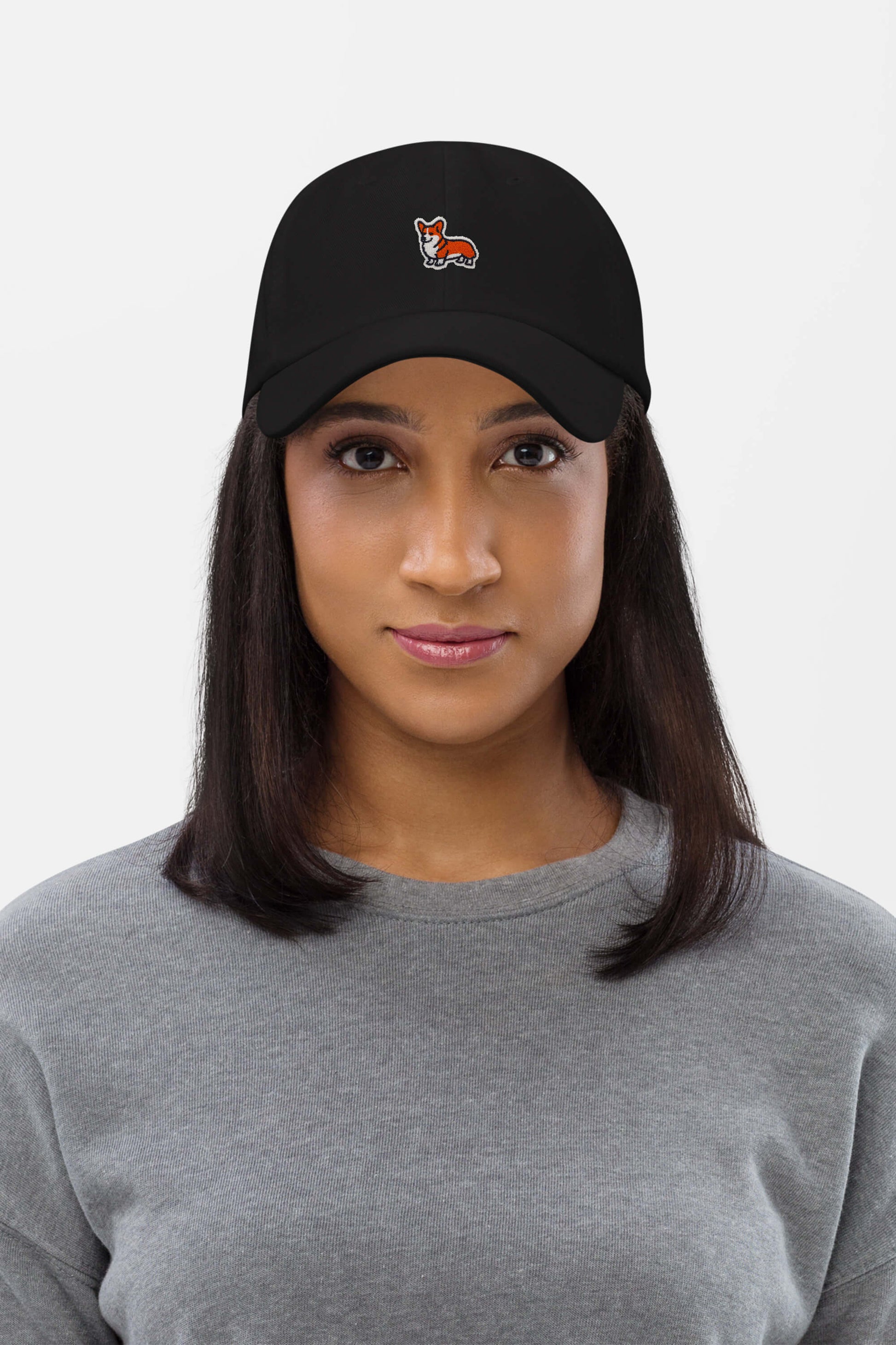 A woman with straight dark hair wears a gray sweatshirt and the Iconic Corgi Cap from Dogloverclothing.com, a black baseball cap featuring a small embroidered corgi on the front, standing against a plain white background.