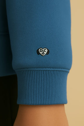 A close-up of the Dog Mom Hoodie by Dogloverclothing.com shows its blue ultra-soft sleeve with a ribbed cuff, a small DS cartoon face patch near the wrist, and subtle paw-print accents.