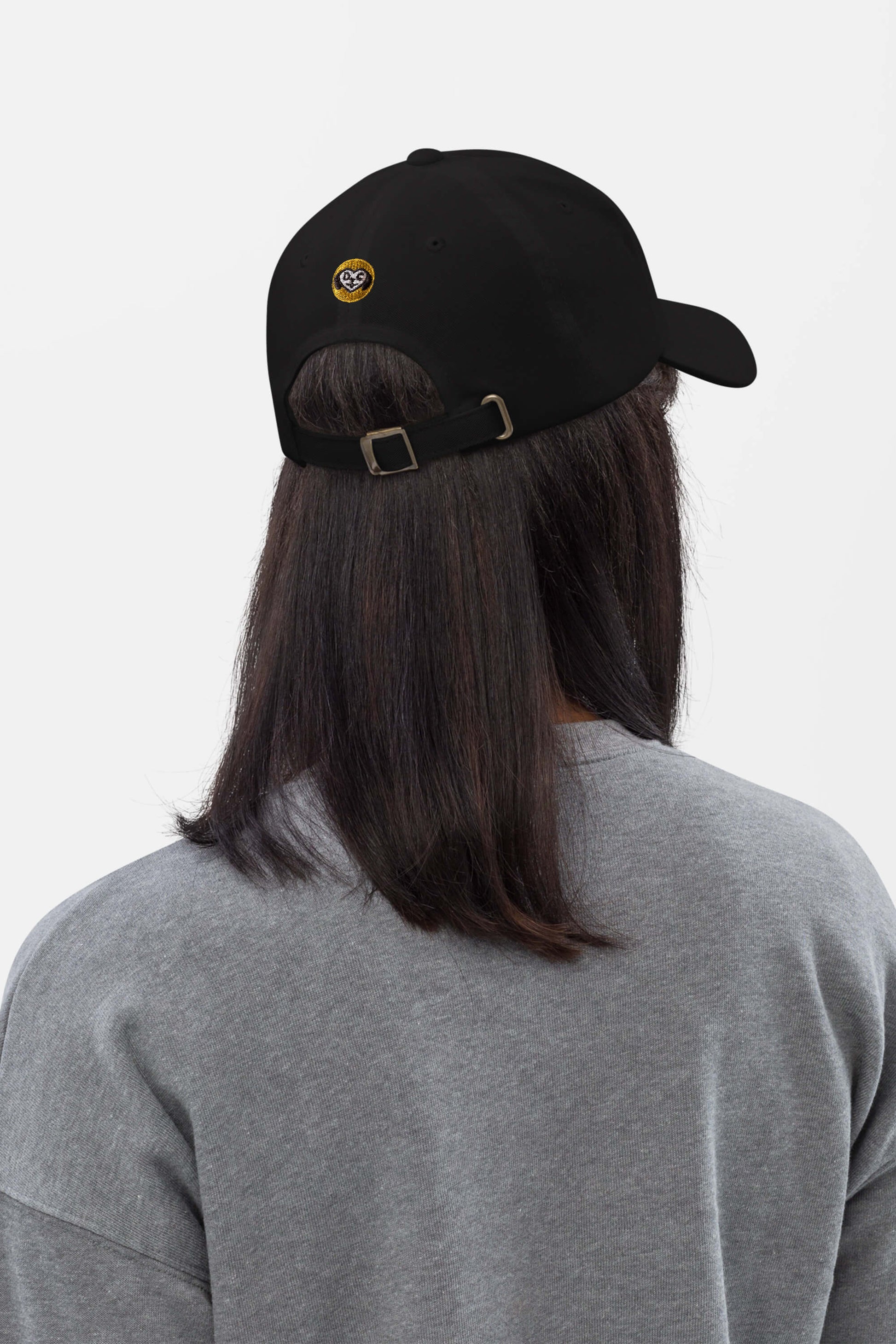 A person with long, dark hair wears Dogloverclothing.coms Iconic Corgi Cap, which features a small yellow and white patch on the back, along with a grey crewneck sweatshirt. The person faces away from the camera against a plain background.