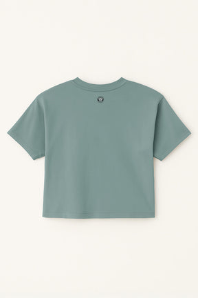An Oversized Dog Mom Crop Top by Dogloverclothing.com is shown laid flat, back side up, in teal with a small circular logo near the neckline on a light cream background.